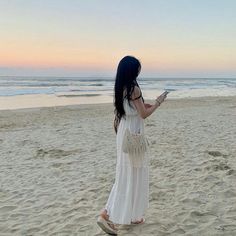 a woman standing on top of a sandy beach next to the ocean holding a cell phone