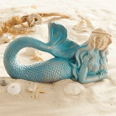 a blue mermaid figurine sitting on top of a sandy beach next to shells