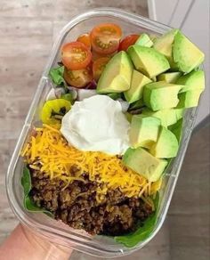 a hand holding a plastic container filled with salad and avocado slices, tomatoes, cheese, sour cream and other toppings