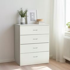 a white dresser with three drawers in front of a window and a potted plant on top