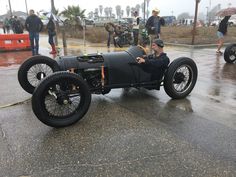 a man driving an old fashioned race car on a wet road with other people in the background