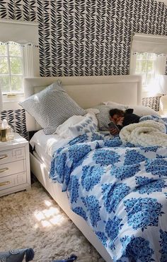 a person laying on a bed with blue and white comforter in a bedroom next to two windows