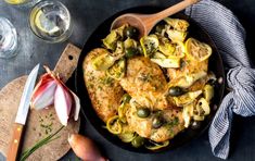 a skillet filled with chicken, artichokes and vegetables next to wine glasses