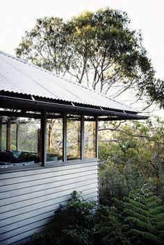 a small white house with a metal roof and windows on the side of it, surrounded by greenery