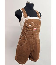 Short overalls with a multi pocket bib and logo patch detail. Traditional straps with clasp closures. Pockets on the front and back with button closures at the sides. Looks great paired with crop tops. SMALL: Waist: 26" | Hips: 40" | Inseam: 3.5" MEDIUM: Waist: 28" | Hip: 42" | Inseam: 3.5" LARGE: Waist: 30" | Hip: 44" | Inseam: 3.75" Material | 100% Cotton (Non-Stretchy Canvas/Denim) Care Instructions | Machine Wash or Hand Wash Cold  Measurements | 30" or 76 cm Top to Bottom 3.5" or 9 cm Inseam [Measured on a size Small] Model is 5'8 and wearing a size Small SMALL CUSTOMER REVIEWS 1. I'm 5'4 about 120 pounds and I ordered a small in the navy. They fit perfectly! I would recommend getting the size you usually wear. The fabric and stitching feel high quality. 2. The overalls are just great Overalls Tank Top Outfit, Brown Cotton Overalls With Pockets, Brown Cotton Overalls, Utility Style Overalls With Bib Front And Suspenders, Utility Style Overalls With Suspenders And Bib Front, Utility Overalls With Suspenders And Bib Front, Utility Bib Front Overalls With Suspenders, Utility Overalls With Button Closure, Utility Shortalls With Suspenders