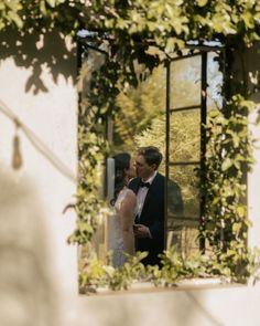 a bride and groom standing in front of a window with greenery on the outside