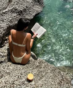 a woman reading a book while sitting on the edge of a cliff next to a body of water