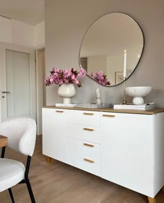 a white dresser with flowers on top and a round mirror above it in a room