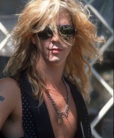 a woman with long blonde hair wearing sunglasses and a black top is standing in front of a chain link fence