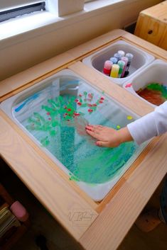 a child is playing with water in a tray on the table next to a window