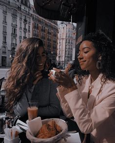 two women sitting at an outdoor table with drinks in their hands and one holding a cell phone to her ear