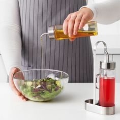 a person pouring dressing into a bowl with salad in the foreground and a blender behind them