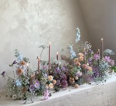 an arrangement of flowers and candles on a white tablecloth with wall in the background