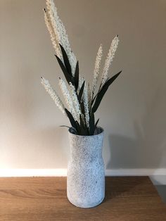 a white vase filled with lots of flowers on top of a wooden table next to a wall