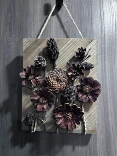 a wooden sign with flowers and pine cones hanging on a rope against a wood wall