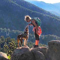 a woman petting a dog on the top of a rock in front of mountains