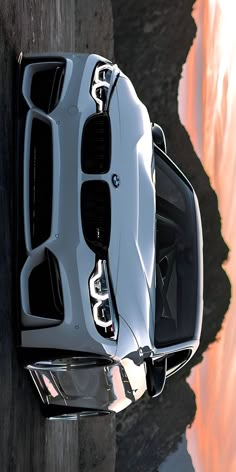 the front end of a white sports car parked next to a body of water at sunset