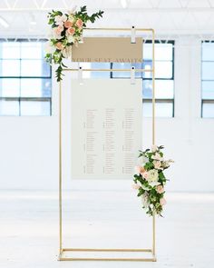 a white and gold wedding seating sign with flowers on the front, along with greenery