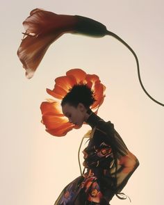 a woman with an orange flower on her head and hair blowing in the wind, against a gray sky