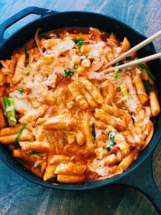 a skillet filled with pasta and vegetables on top of a wooden table next to chopsticks