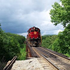 a red train traveling over a bridge next to lush green trees on a cloudy day