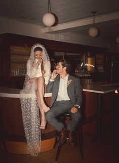 a man sitting next to a woman on top of a stool in a bar wearing a wedding dress