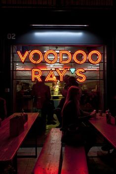 people sitting at wooden benches in front of a neon sign that says voodoo rays on it