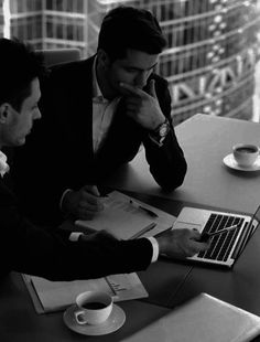 two men sitting at a table with laptops and papers