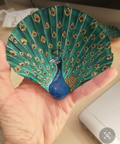 a hand holding a green and blue peacock ornament in front of a computer