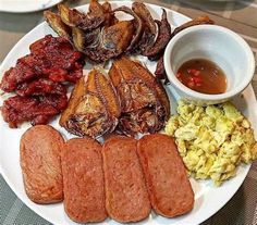 a white plate topped with meats, eggs and other foods next to a bowl of sauce