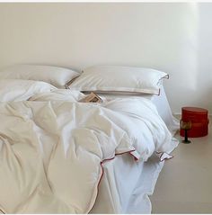 an unmade bed with white sheets, pillows and a red lamp next to it