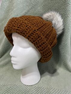 a white mannequin head wearing a brown knitted hat with a grey fur pom - pom