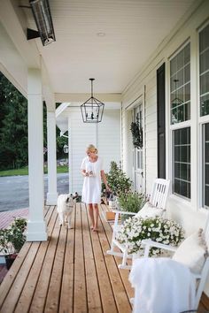 a woman is walking her dog on the porch with white furniture and flowers in pots