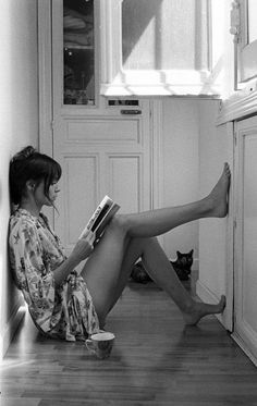 a woman sitting on the floor with her legs crossed and reading a book next to a cat