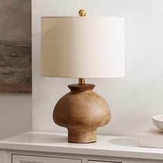 a wooden table lamp sitting on top of a white dresser next to a bowl and painting