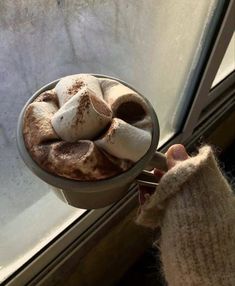 someone is holding a cup of hot chocolate with marshmallows in it and looking out the window