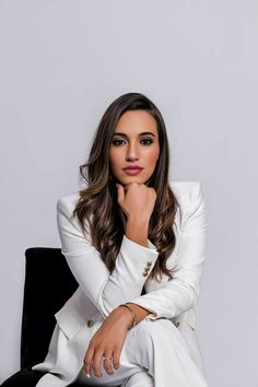 a woman sitting on top of a black chair in front of a white wall with her hand under her chin