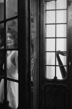 black and white photograph of woman looking out the window at another person in dress standing behind her