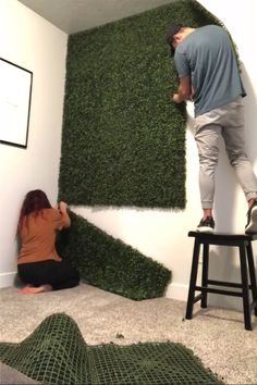 two people are working on a fake grass wall in the corner of a living room