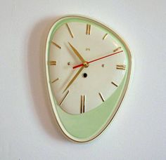 a green and white clock hanging from the side of a wall with red hands on it