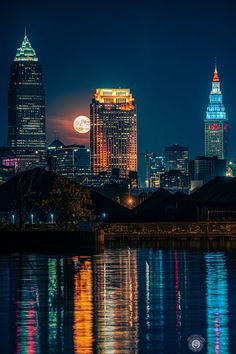 the full moon is reflected in the water next to some tall buildings at night time