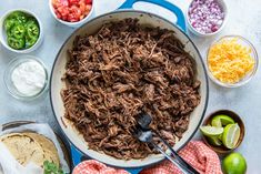 shredded beef in a skillet surrounded by bowls of salsa, tortillas and limes