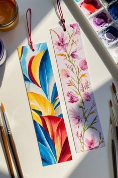 two colorful bookmarks sitting on top of a table next to paint brushes and watercolors