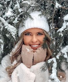 a woman wearing a white hat and scarf in front of snow covered pine trees with her hands on her face