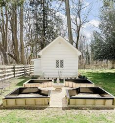 several raised beds in front of a small white house with a shed behind them and trees