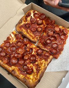 two slices of pepperoni pizza in a box with someone's hand holding a cell phone