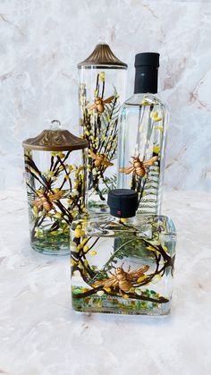 three glass bottles with flowers and bees on them sitting on a marble counter top next to each other