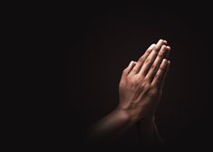 two hands folded in prayer on a dark background with one hand raised up to the sky