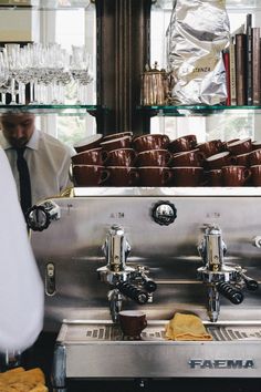 a man standing in front of a coffee machine filled with cups and saucers next to shelves full of books