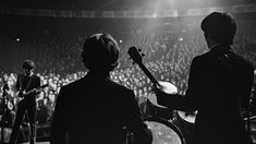 two men are playing instruments in front of an audience at a rock concert, while one man is on the drums